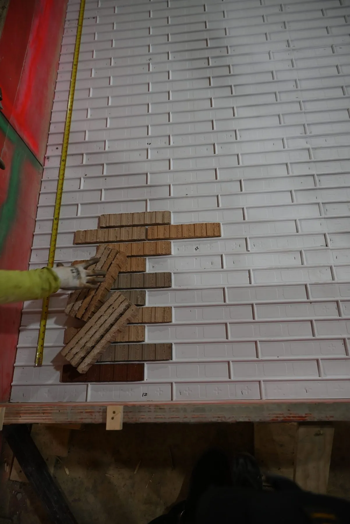 A gloved hand setting clay thin brick face-down into a white rubber form liner on a casting bed, with a tape measure running along the edge