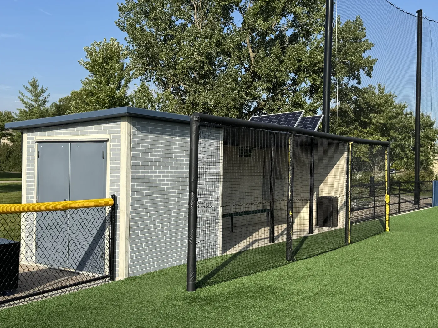 A light blue brick-pattern precast dugout with a dark flat roof beside a batting cage and synthetic turf at Field 32 in Lemont, Illinois