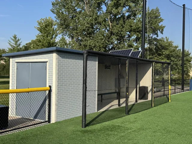 A light blue brick-pattern precast dugout with a dark flat roof beside a batting cage and synthetic turf at Field 32 in Lemont, Illinois