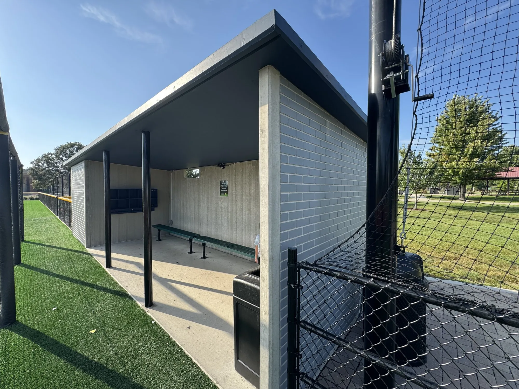 Open front of a Field 32 dugout from the field, the roof carried on steel posts, bench and cubbies along the back wall