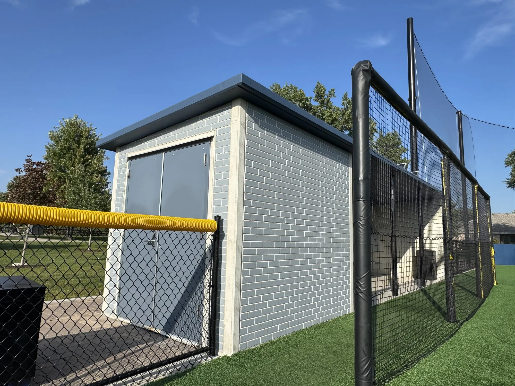 The storage end of a Field 32 dugout, steel double doors in the light blue precast wall, with the fence and backstop netting beyond